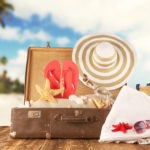 Travel concept with old suitcase on wooden planks full of beach accessories. Placed on mole with sandy beach on background